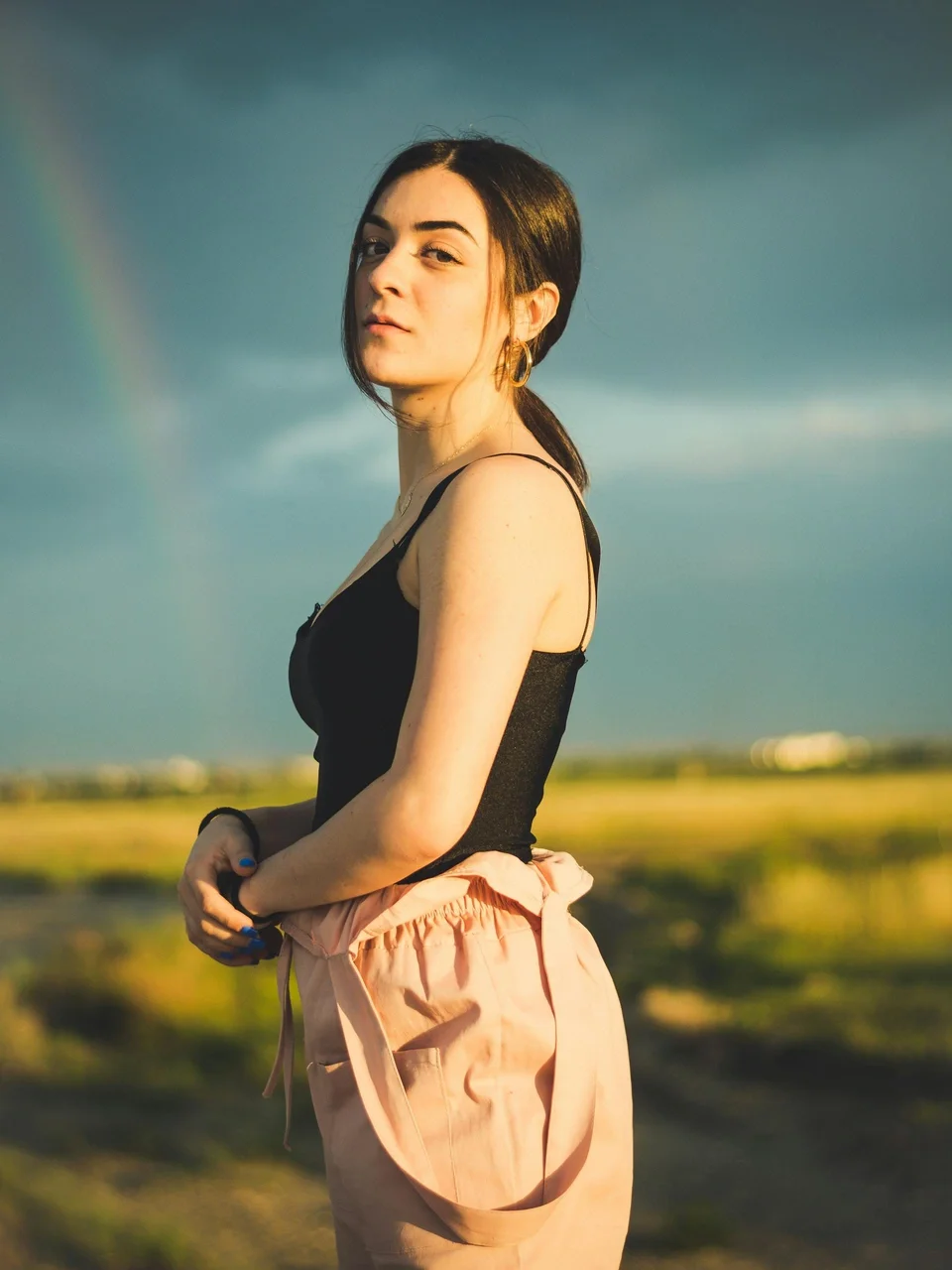 Woman standing confidently in a field at sunset with a rainbow in the background