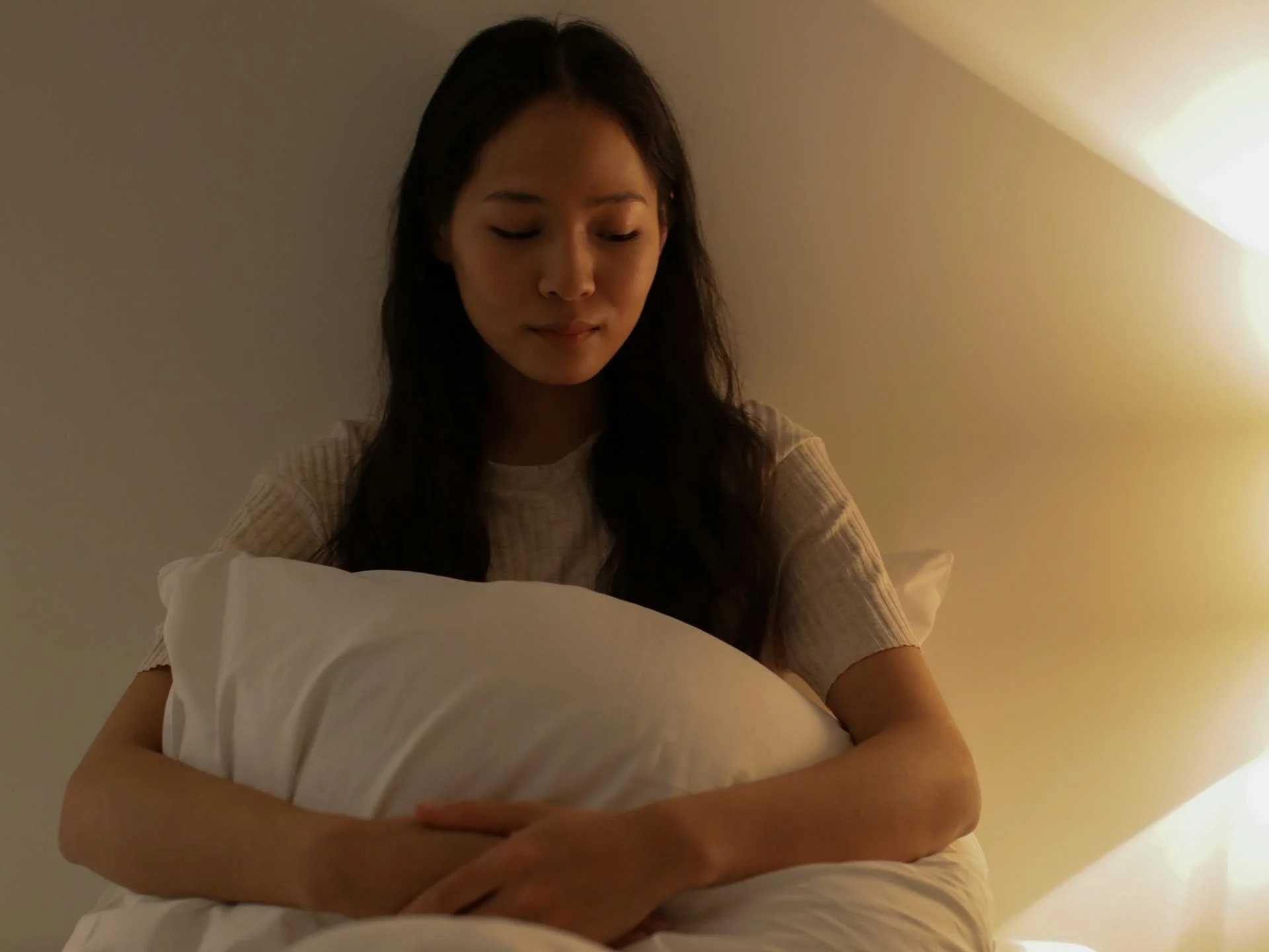 Young woman with closed eyes holding a pillow in a softly lit bedroom